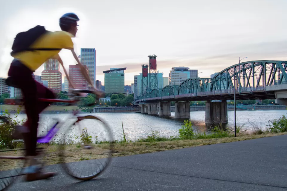 Cyclist on bike path in Portland, Oregon with bridge over river and city skyline in background.